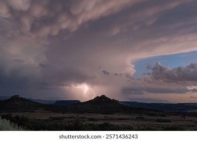 A forked bolt of lightning strikes from a dramatic sky filled with pink and grey clouds above the desert landscape of Hop Valley in Southern Utah, USA on a summer evening.