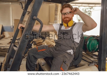 Forklift driver in protective eyeglasses sitting in vehicle in wood warehouse and looks camera