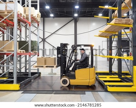 Forklift with drivers. Process of loading pallet onto racks. Multi-tiered racks with boxes and boards. Warehouse worker. Man drives loading truck. Forklift is yellow. Warehouse special equipment