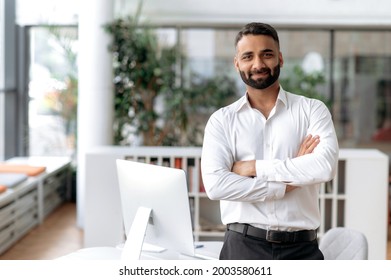 Formal business male portrait. Confident successful Indian businessman or manager, in white shirt, stands near his work desk in the office, arms crossed, looks directly at camera and smiles friendly - Shutterstock ID 2003580611