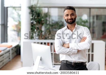 Formal business male portrait. Confident successful Indian businessman or manager, in white shirt, stands near his work desk in the office, arms crossed, looks directly at camera and smiles friendly