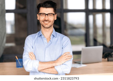 Formal business portrait. Confident successful caucasian businessman or manager with glasses stands near his work desk in the office, arms crossed, looks directly at the camera and smiles friendly - Shutterstock ID 2311823793