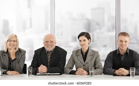 Formal Businessteam Portrait Of Different Generations Sitting At Meeting Table, Smiling At Camera.?