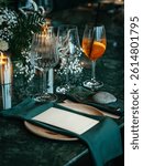 Formal dining table elegantly set with a wine glass, cocktail glass, plates, and green napkins. Florals and candles add a sophisticated touch, perfect for a fine dining experience