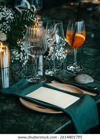  Formal dining table elegantly set with a wine glass, cocktail glass, plates, and green napkins. Florals and candles add a sophisticated touch, perfect for a fine dining experience