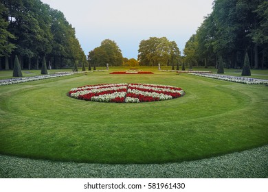 Formal garden with round flower bed surrounded by pyramidal bushes and big green trees.