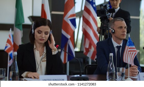 Formal international politicians in headphones sitting at table on summit and listening to speech translation in headphones