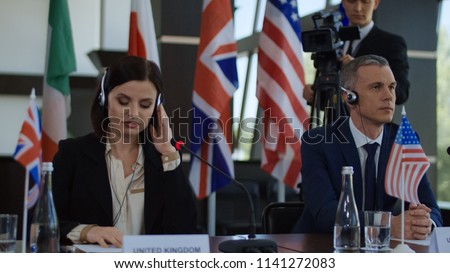 Formal international politicians in headphones sitting at table on summit and listening to speech translation in headphones