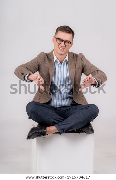 Formal man sitting on cube with legs crossed.\
Male showing both thumbs down, with a disguss to the camera. Gray\
background.