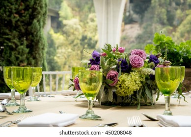 Formal table setting for a wedding with floral centerpiece and colorful green glasses in front of a large view window overlooking a garden