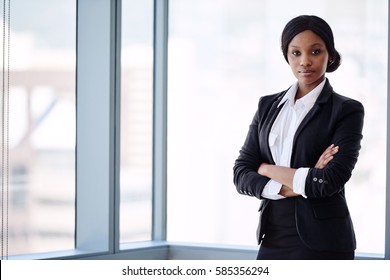 formally dressed african business woman looking into the camera with her arms crossed while standing in front of large glass windows with a cityscape behind her.