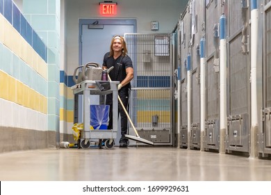 Fort Lauderdale, Florida / USA - 3/28/2020: Young Fit Man Wearing Gloves And A Blue Uniform Cleaning The Floor Of A Dog Kennel With Water Soap And A Squeegee At A County Animal Shelter Near Cage Door