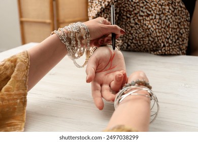Fortune teller reading lines on woman's palm at white wooden table, closeup. Chiromancy