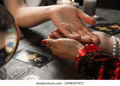 Fortune teller reading lines on woman's palm at black table, closeup. Chiromancy