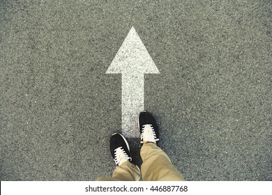 Forward Arrow Painted On An Asphalt Road. Top View Of The Legs And Shoes. POV