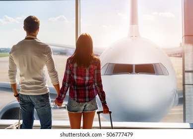 Forward To Future. Young Loving Couple Is Watching Plane Though Window Before Departure. They Are Standing At Airport And Holding Hands