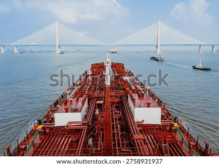 Forward view from a massive cargo tanker deck with complex red piping towards a long modern cable-stayed bridge and other vessels on a river waterway