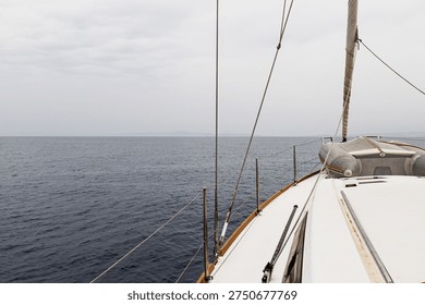 A forward-facing view from the deck of a sailboat navigating on calm open water. The rigging and part of the mast are visible, along with an inflatable dinghy secured near the bow. The distant horizon and soft outlines of land create a peaceful maritime atmosphere. The image highlights sailing lifestyle, nautical travel, and the experience of being at sea.