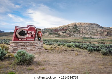 Fossil Butte National Monument sign in front of painted red hills in sagebrush desert.