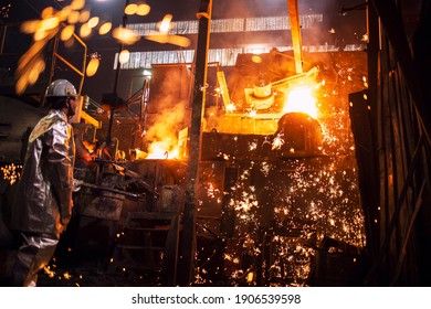 Foundry worker controlling process of melting iron in furnace. Burning liquid steel pouring and sparks flying all around. Metallurgy and heavy industry.