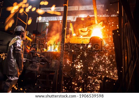 Foundry worker controlling process of melting iron in furnace. Burning liquid steel pouring and sparks flying all around. Metallurgy and heavy industry.