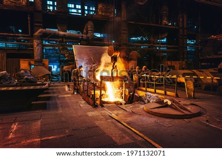 Foundry workshop interior, molten iron pouring from blast furnace into ladle container and workers founders control process, heavy metallurgy industry