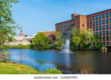 Fountain On The Nashua River Against The Background Of A Historic Cotton Factory Building With A Clock Tower In The Old Industrial Park Of Nashua. New Hampshire, USA