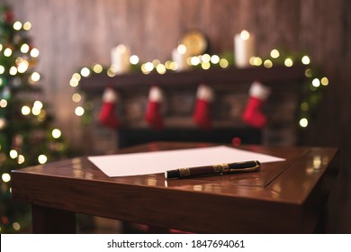 Fountain Pen, Sheet Of Paper On The Table On Christmas Tree And Fireplace With Decoration Of Light Bulbs Background. Concept Of Writing Letter To Santa Claus 