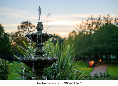 Fountain with water drops in a park with trees behind at dusk. The fountain is seen with a path, bench and lights just after sunset. This fountain is a central feature in the park. - Shutterstock ID 2057037674