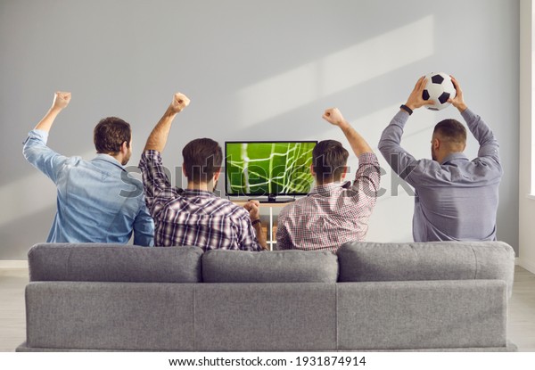 Four adult male friends with arms raised\
emotionally watching a football match on TV at home. Unrecognizable\
friends sit on the couch with their backs to the camera and cheer\
for their favorite team.