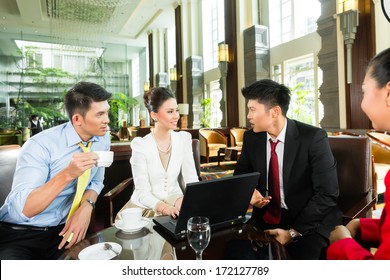 Four Asian Chinese Office People Or Businessmen And Businesswomen Having A Business Meeting In A Hotel Lobby Discussing Documents On A Tablet Computer While Drinking Coffee 