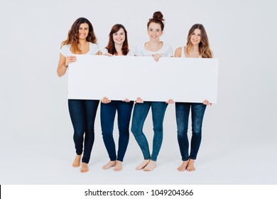 Four Attractive Barefoot Women In Jeans Standing In A Line Holding A Blank White Rectangular Panoramic Sign With Copy Space Isolated On White