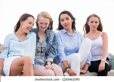 Four Attractive Women In Summer Clothes Sitting At Concrete Border Near River After Studing In College. Young Student Girls Having Fun