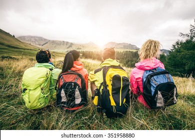 Four Backpackers Looking At Sunset Over The Mountains - Hikers Talking And Relaxing After An Excursion In The Nature - Friends Enjoying Winter Holiday