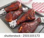 Four barbequed turkey legs, wrapped in foil on one end for handling, sit on a metal baking dish ready for eating