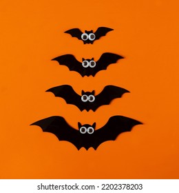 Four Black Bats With White Eyes Aligned Verticaly On Orange Background. Spooky Minimal Creative Concept For Halloween Wallpaper Or Web Banner Or Advertisement. Flat Lay