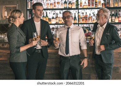 Four Business Partner Celebrating Their Victory. Old Business Man Making A Toss To Younger Business Man And Woman. They Are Holding A Champagne Glass In A Hotel Lobby With City Scape Background.