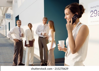 Four Business People Talking In Corridor Outside Meeting Room, Focus On Businesswoman Using Mobile Phone, Smiling