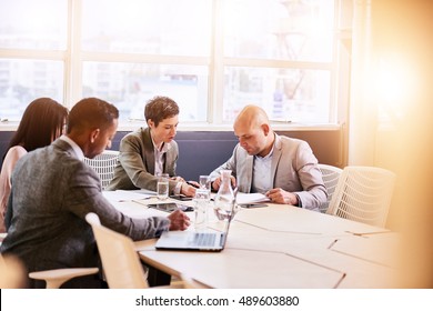 Four Business Professionals Conducting A Meeting In A Bright Modern Conference Room With A Large Windown And An Abundance Of Natural Light, While Making Use Of Technology To Optimise Efficiency.