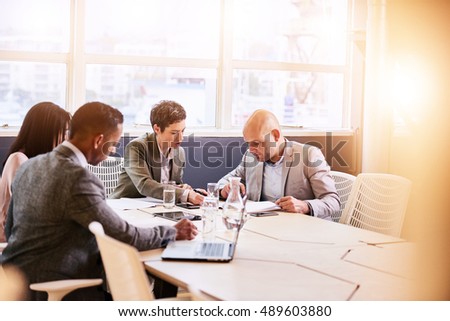 Four business professionals conducting a meeting in a bright modern conference room with a large windown and an abundance of natural light, while making use of technology to optimise efficiency.