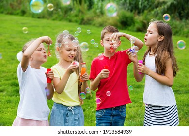 Four Cheerful Kids In School Age Sitting Together In Park And Blowing Soap Bubbles