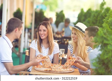 Four Cheerful Young Friends Sharing Pizza In A Outdoor Cafe