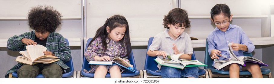 Four Children Reading On A Chair In The Classroom, Young Students Study In The Library. Education Home School Back To School Concept Background Panoramic Banner.