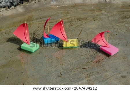 Four children's colorful ships are floating in a shallow pond. The toy sailboats are made from kitchen sponges. Summer, daytime. Selective focus. No people.