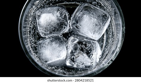 Four clear ice cubes float in a glass of bubbly, clear liquid, likely soda water or tonic, against a dark background.