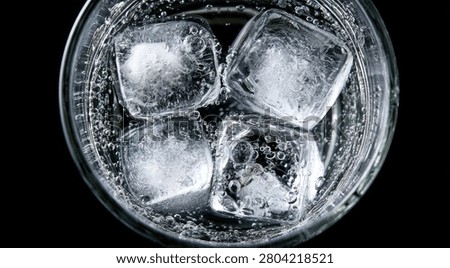Four clear ice cubes float in a glass of bubbly, clear liquid, likely soda water or tonic, against a dark background.