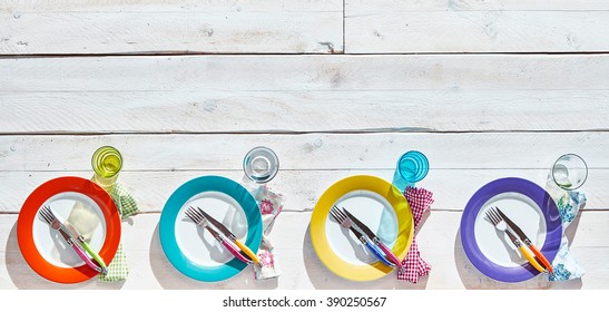 Four colorful empty plates with matching forks, knives and napkins on painted white wooden table with copy space above - Powered by Shutterstock