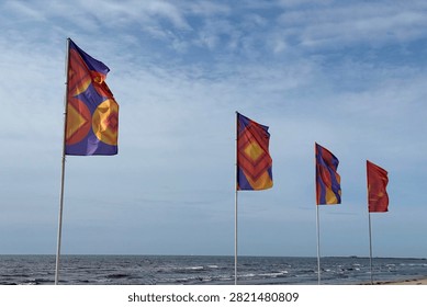 Four colorful patterned flags wave along a sandy coast under a cloudy summer sky. The horizontal low-angle shot captures geometric designs, sea views, outdoor festival decor, and beach energy.