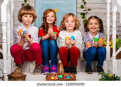Four cute children, two boys and two girls are sitting next to each other on a wooden threshold with Easter eggs in their hands. Easter