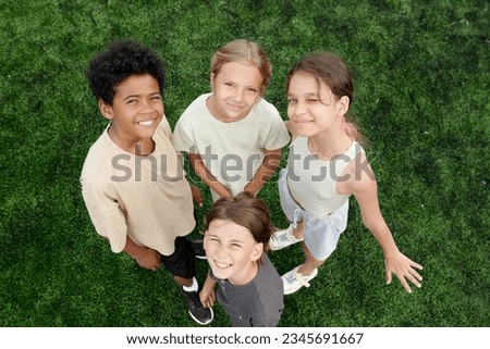 Four cute happy schoolchildren in activewear looking at camera while standing close to one another on green lawn or football field